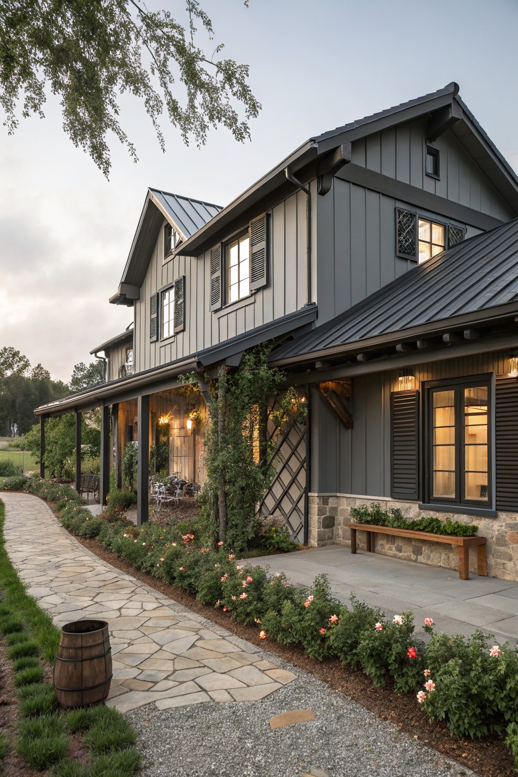 Side exterior of a two-story modern farmhouse with dark gray board-and-batten siding, black metal roof, covered porch with wooden posts, stone foundation, shuttered windows, and a flagstone path with plants and a wooden barrel planter.