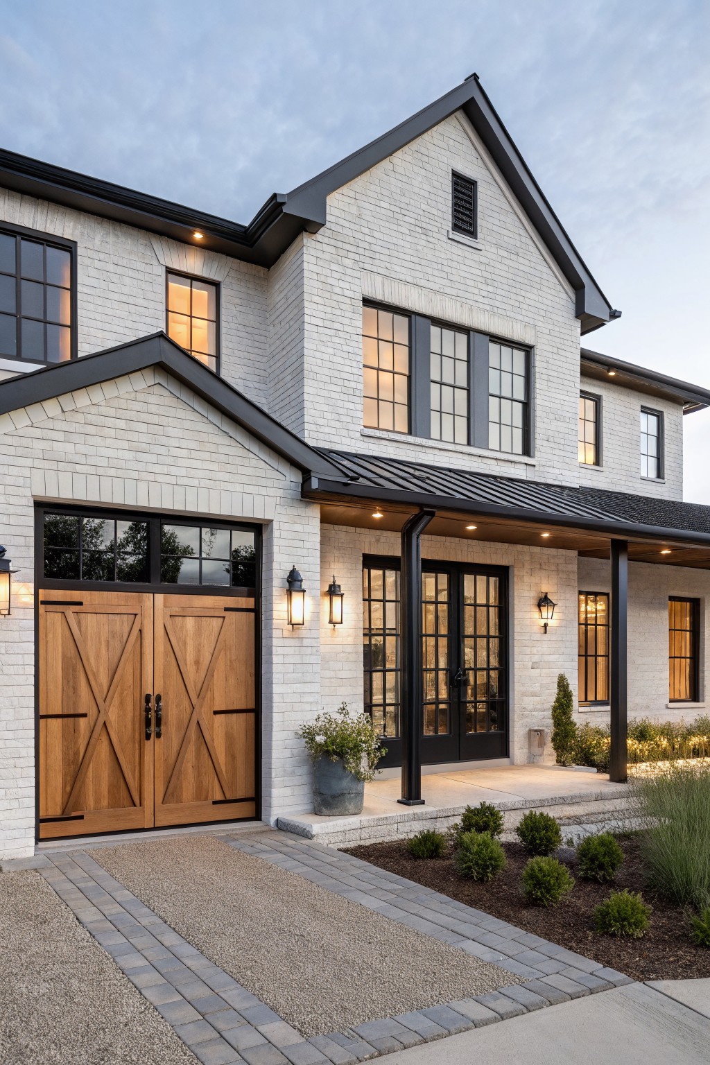 White brick modern farmhouse exterior with black-framed windows, wooden garage doors, covered front porch, and gravel pathway at dusk.