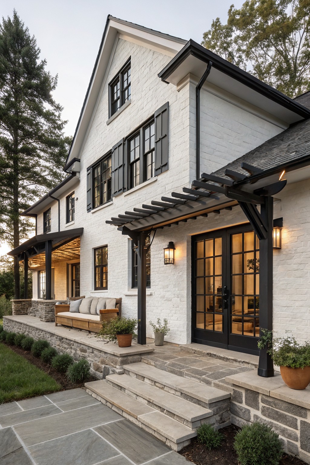 Side exterior of a two-story white brick house with black-framed multi-pane windows, black shutters, black double glass doors under a black pergola, stone steps leading up from a bluestone patio, and low plantings in a yard with trees.