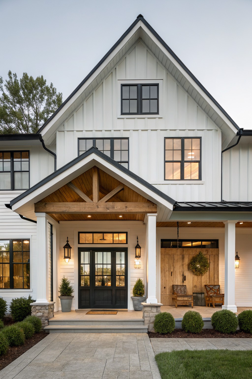 White board-and-batten sided two-story modern farmhouse exterior with black-framed windows and doors, wooden-beamed covered porch, lanterns, potted trees, boxwoods, and stone steps at dusk.