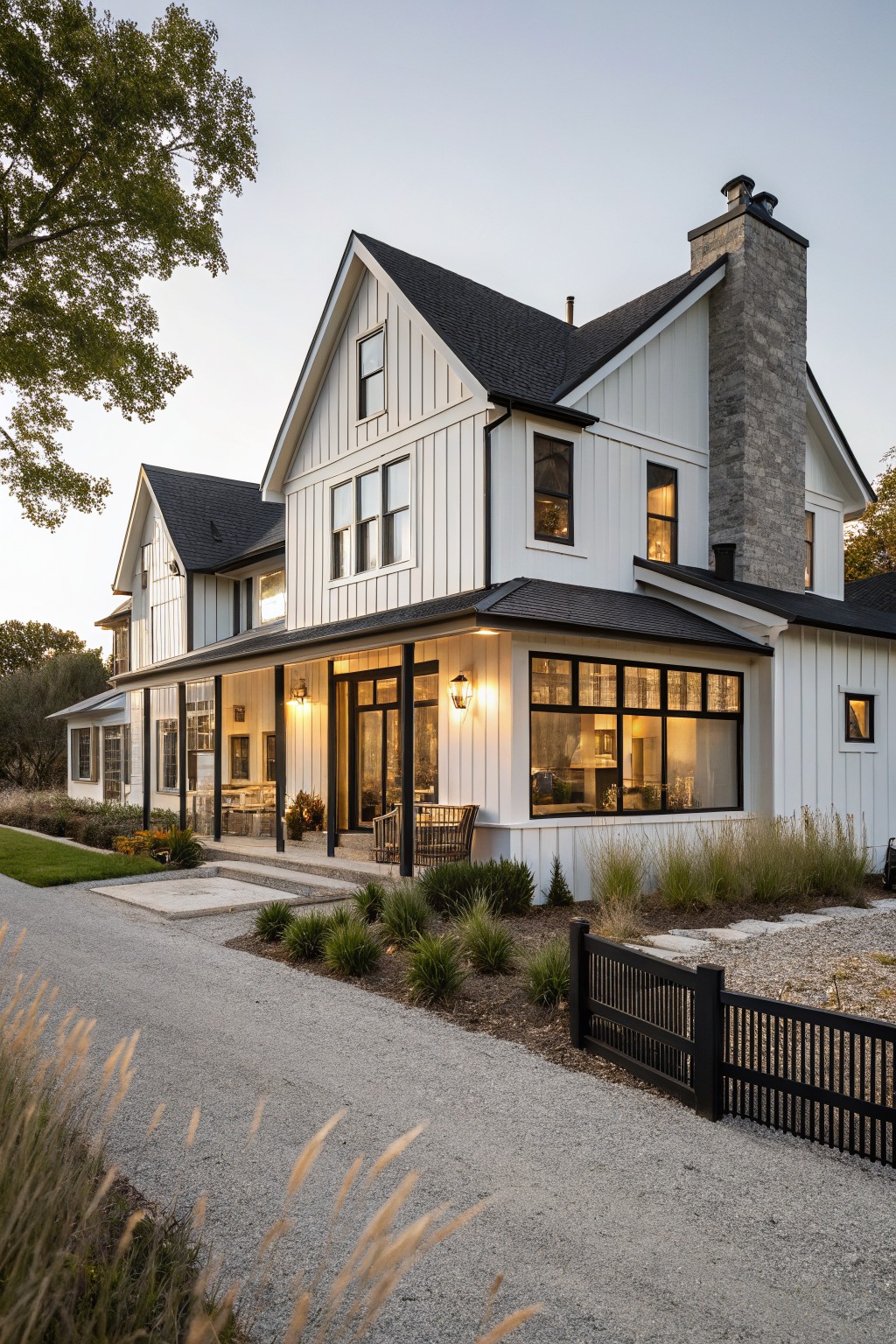 A two-story white board-and-batten modern farmhouse exterior with black-framed windows, black roof sections, stone chimney, wrap-around porch, gravel driveway, and ornamental grasses at dusk.