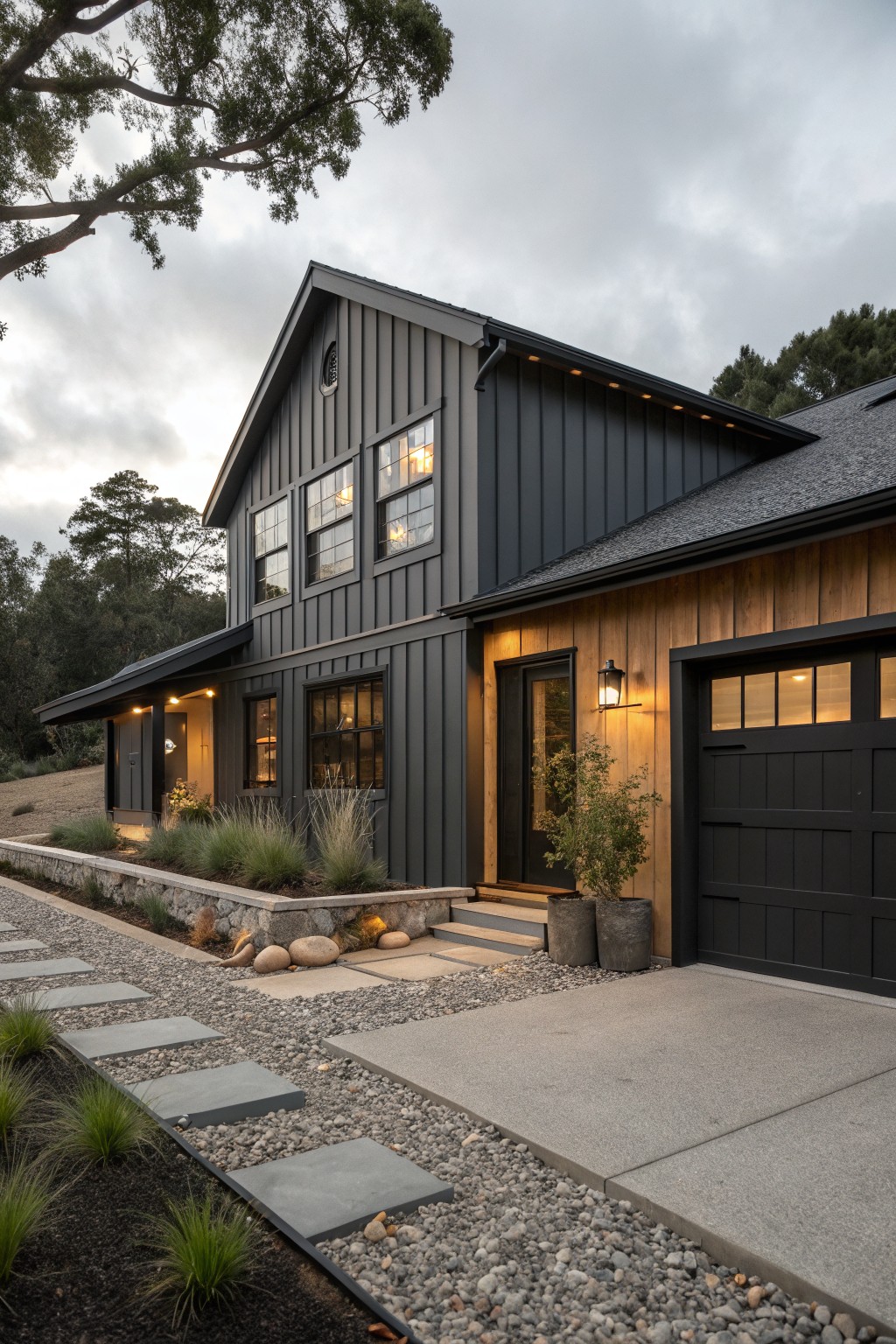 Two-story modern farmhouse house exterior with dark gray board-and-batten siding, multiple black-framed windows, wood-paneled garage door, entry with lanterns, stone pathway, gravel driveway, and low native grasses under cloudy sky.