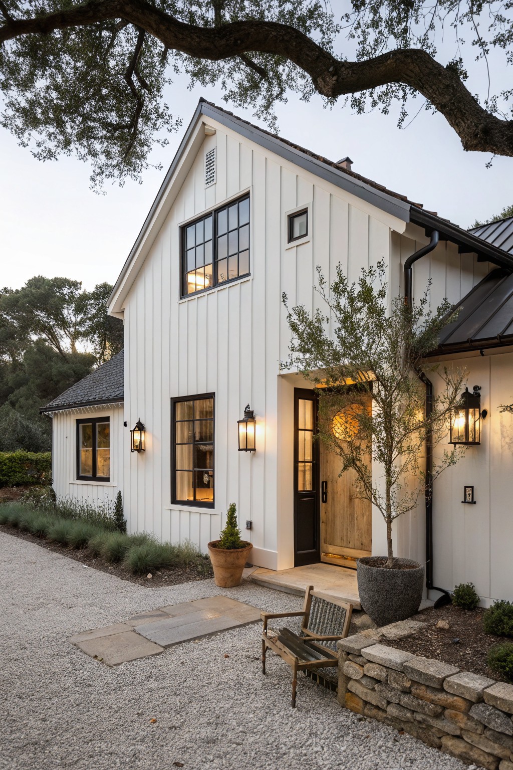White board-and-batten sided modern farmhouse exterior with black-framed windows, dark metal roof, wooden entry door, lanterns, potted olive tree, gravel driveway, stone wall, and low shrubs under oak trees at dusk.