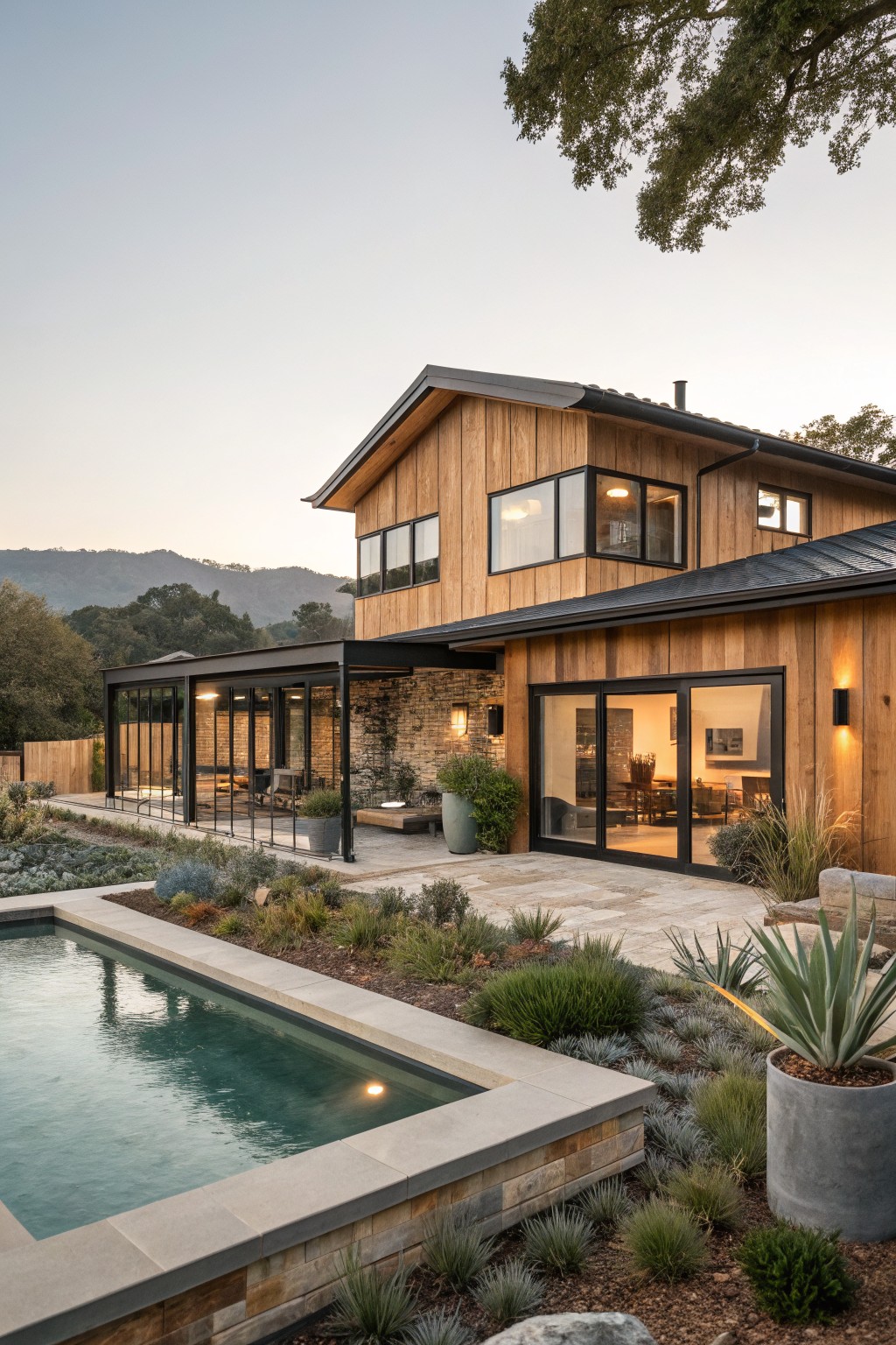 Two-story house exterior with vertical wood siding, black-framed windows, dark metal roof, glass-walled patio, stone accents, infinity-edge pool, and drought-tolerant landscaping on a hillside at dusk.