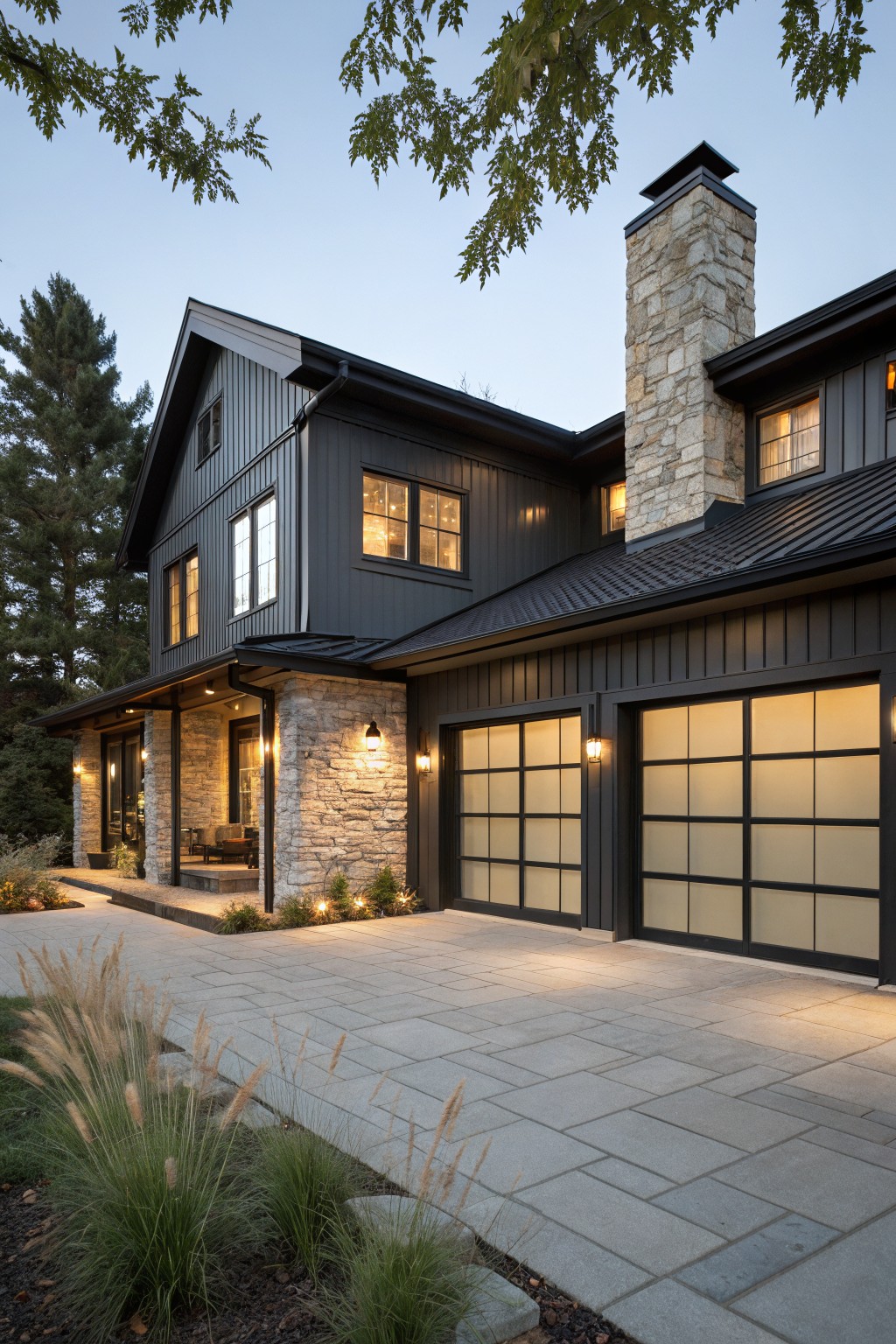 Exterior of a two-story modern farmhouse home with black board-and-batten siding, stone chimney and accents, covered porch, double garage doors with black frames and frosted glass grids, pathway lighting, and ornamental grasses along a paver driveway.