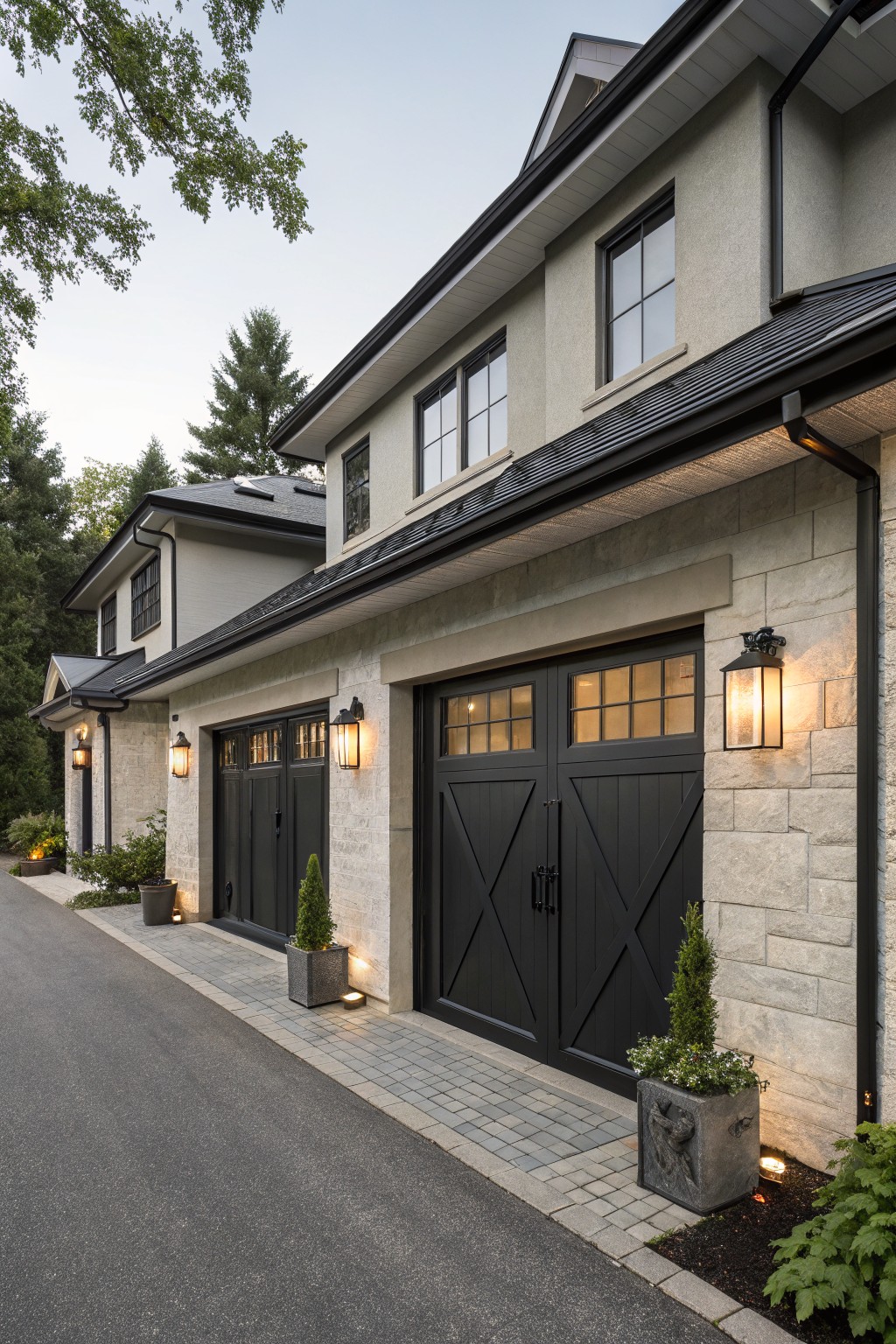 Two-car garage on a modern house exterior with black paneled wooden doors featuring glass windows and X bracing, light beige stone side walls, wall lanterns, and potted plants along a paved driveway edged in pavers.