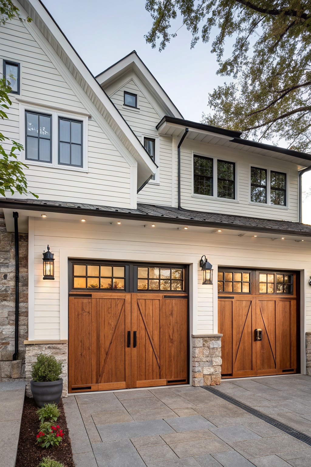 Modern farmhouse house exterior with two dark wood carriage-style garage doors featuring X-bracing, glass panels, and black hardware, set against white shiplap siding, black-framed windows, stone pillars, lantern lights, and a gray paver driveway with edging plants.