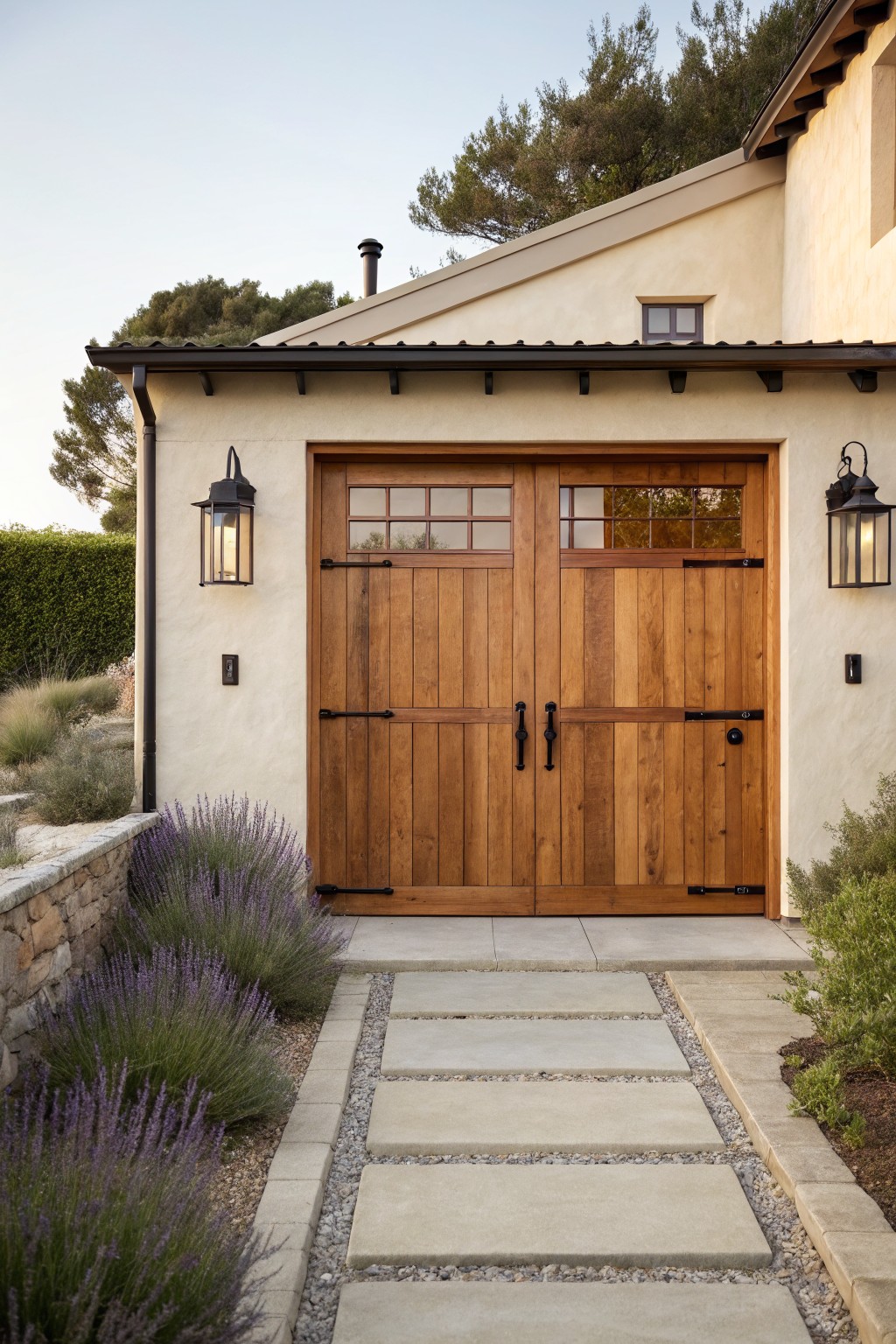 Double wooden plank garage doors with divided glass windows and black wrought-iron strap hinges and handles on a beige stucco wall, flanked by black lanterns, beside a concrete paver pathway edged with lavender shrubs and gravel.