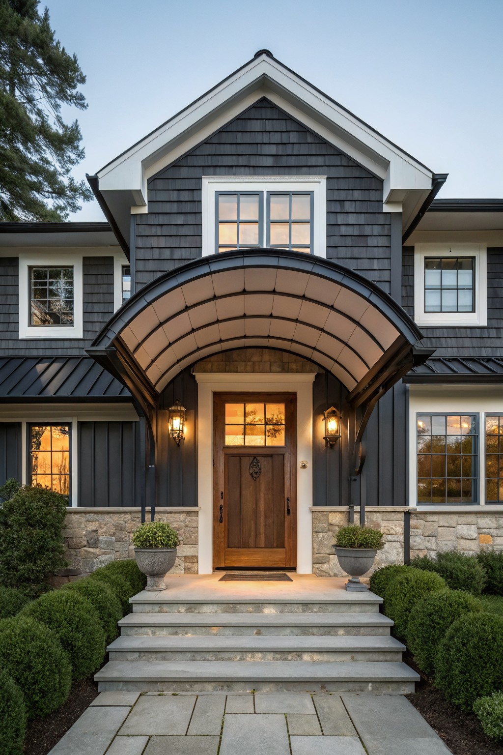 Front view of a two-story modern farmhouse house with dark shingle siding, white trim, gabled roof, arched white canopy over a wooden entry door with lanterns, stone pillars, potted plants, boxwoods, and stone steps and path.