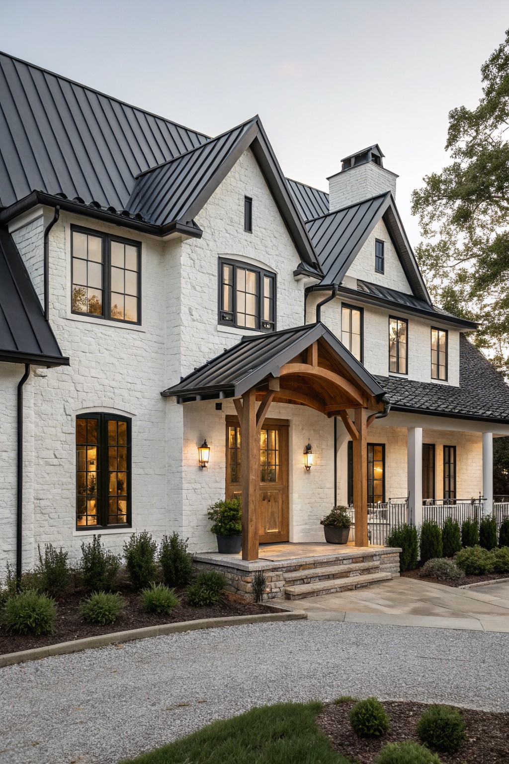 Two-story modern farmhouse exterior with white brick walls, black standing seam metal gabled roofs, wooden beam portico over front door, black-framed windows, lanterns, shrubs, and gravel driveway.