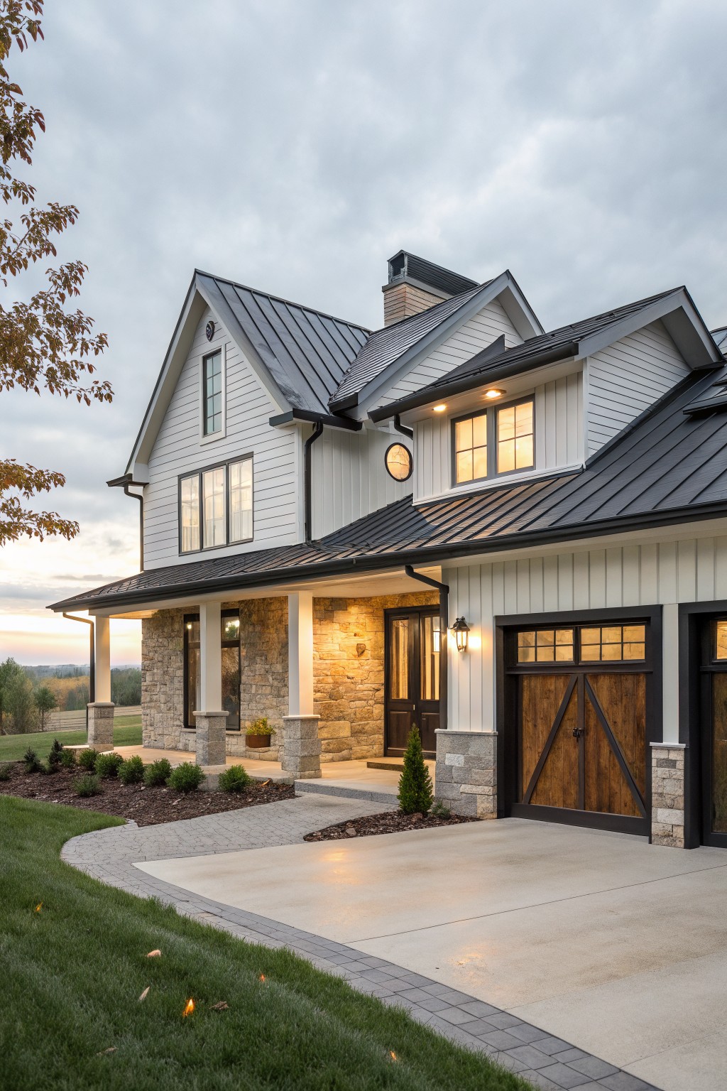 Side exterior view of a two-story modern farmhouse with white board-and-batten siding, black standing seam metal roof, stone pillar porch, wooden garage doors, and landscaped yard at dusk.