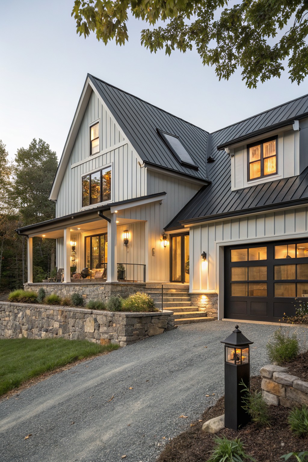 Two-story modern farmhouse with light gray vertical board-and-batten siding, dark standing seam metal gable roof, wraparound porch, attached garage, stone retaining walls, gravel driveway, and landscaping at dusk.