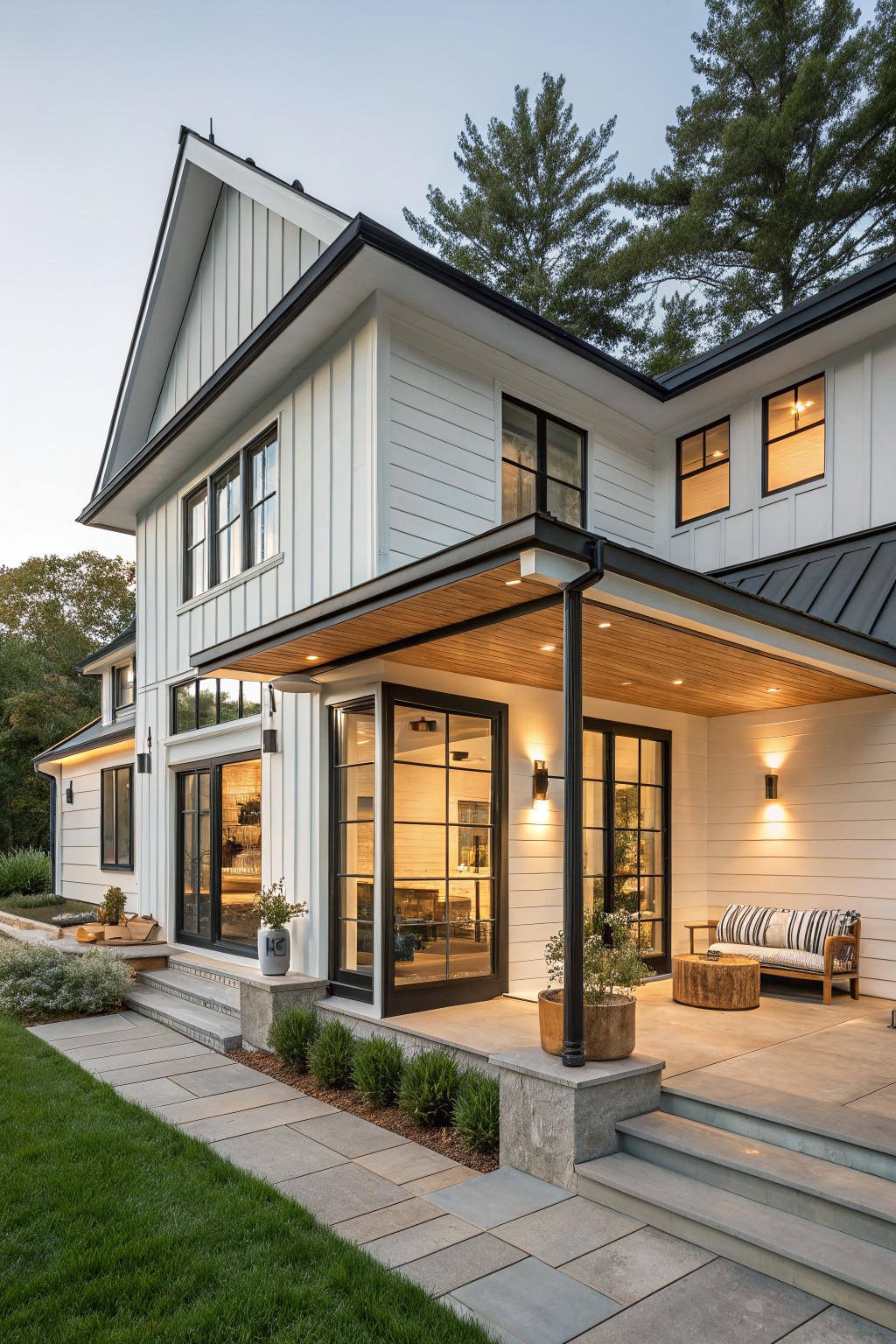 Side exterior view of a two-story modern farmhouse with white vertical board-and-batten siding, dark standing seam metal gable roof extending over a covered porch, black-framed windows and doors, stone steps, and a bluestone path bordered by low shrubs and grass.
