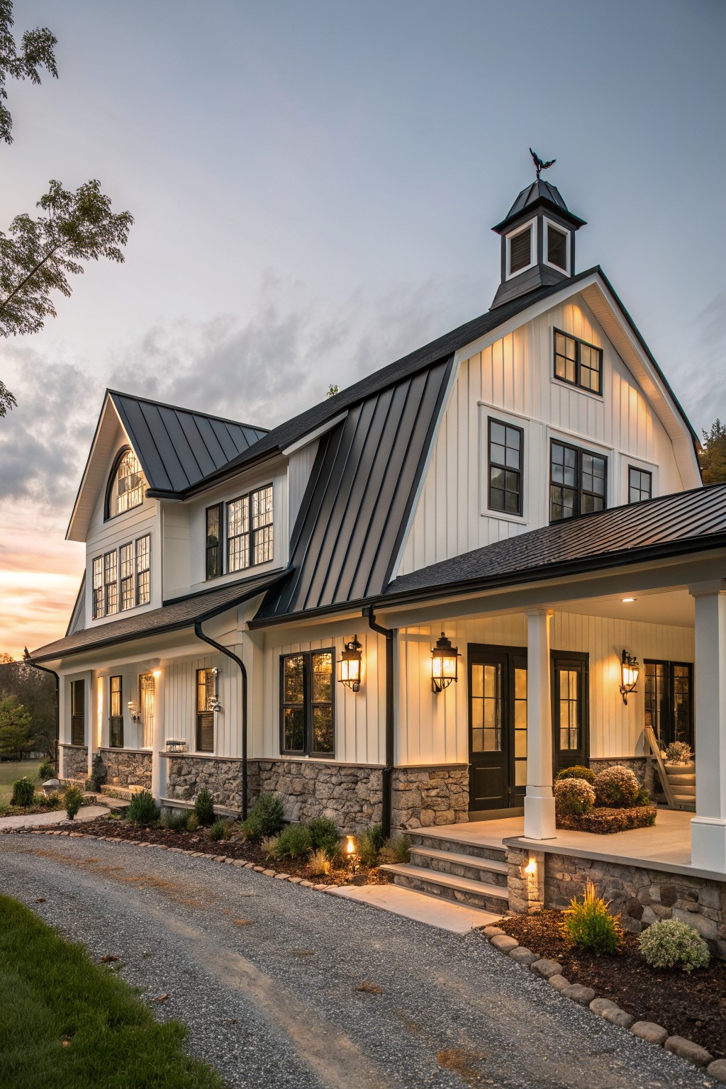Two-story modern farmhouse exterior with dark standing seam metal roof, white board-and-batten siding, black-framed windows, cupola with weathervane, covered porch with lanterns, stone foundation, and gravel driveway at dusk.