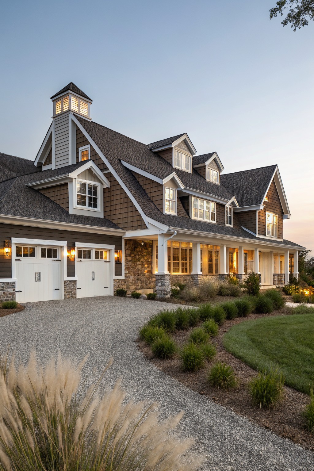 Two-story modern farmhouse house exterior at dusk with dark shingled multi-gabled roof, dormer windows, cupola lantern, white garage doors, stone base, wraparound porch, gravel driveway, and ornamental grasses.