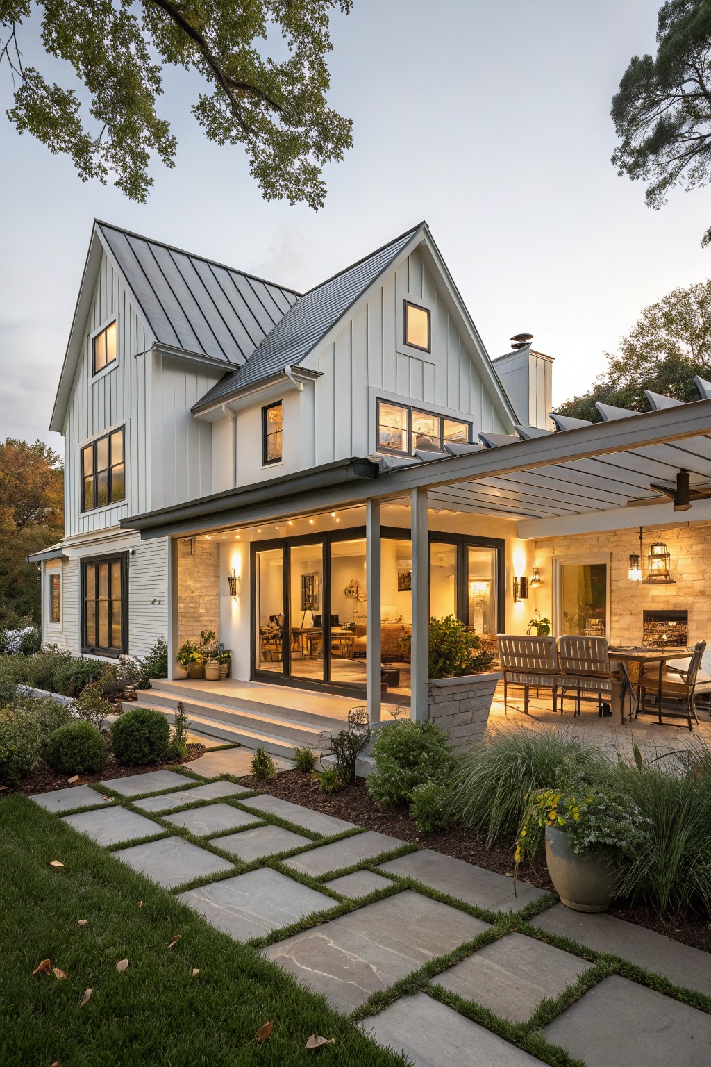 Two-story modern farmhouse exterior with white board-and-batten siding, gray standing seam metal gabled roof, covered porch with pergola and ceiling fan, stone chimney and accents, glass doors opening to patio dining area, and concrete path through landscaped yard with grasses and shrubs.