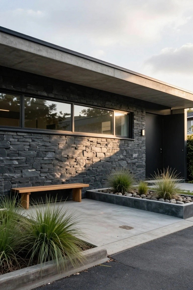 Modern house exterior with dark irregular stone wall cladding beside a black entry door, large reflective glass window, concrete overhang, wooden bench, raised bed of grasses and pebbles, and concrete path next to asphalt driveway.