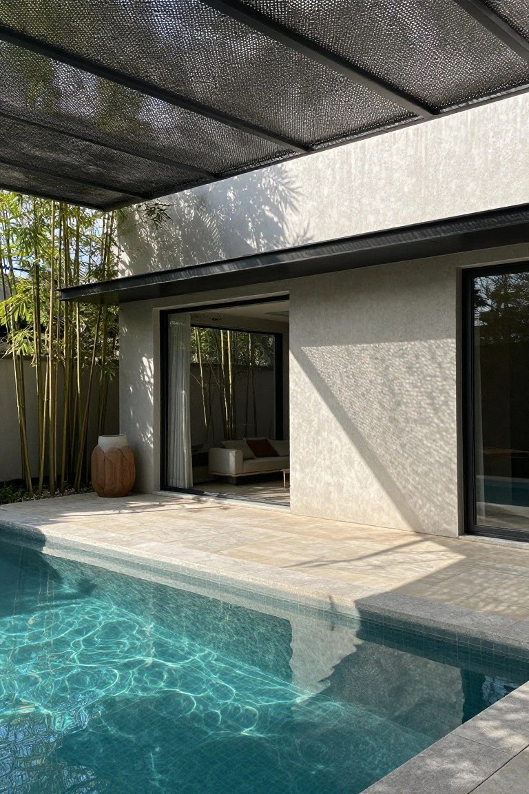 Modern outdoor pool area with turquoise tiled water adjacent to a beige stone terrace, open floor-to-ceiling sliding glass doors on a light textured plaster house wall revealing an indoor white sofa with cushions, tall green bamboo plants along one side, a large carved wooden pot, black metal framed windows and doors, and a black mesh shade canopy overhead.