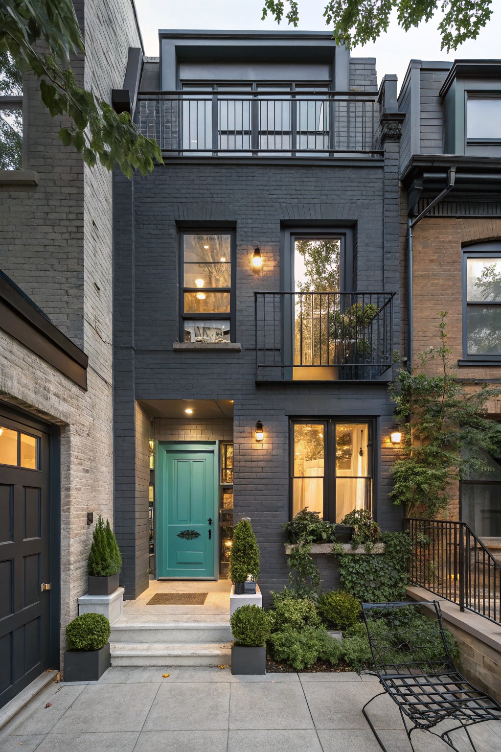 Three-story attached brick house with dark gray painted brick facade, turquoise front door with brass knocker, black metal balcony above entry, potted shrubs flanking steps, and adjacent brick neighbors in evening light.