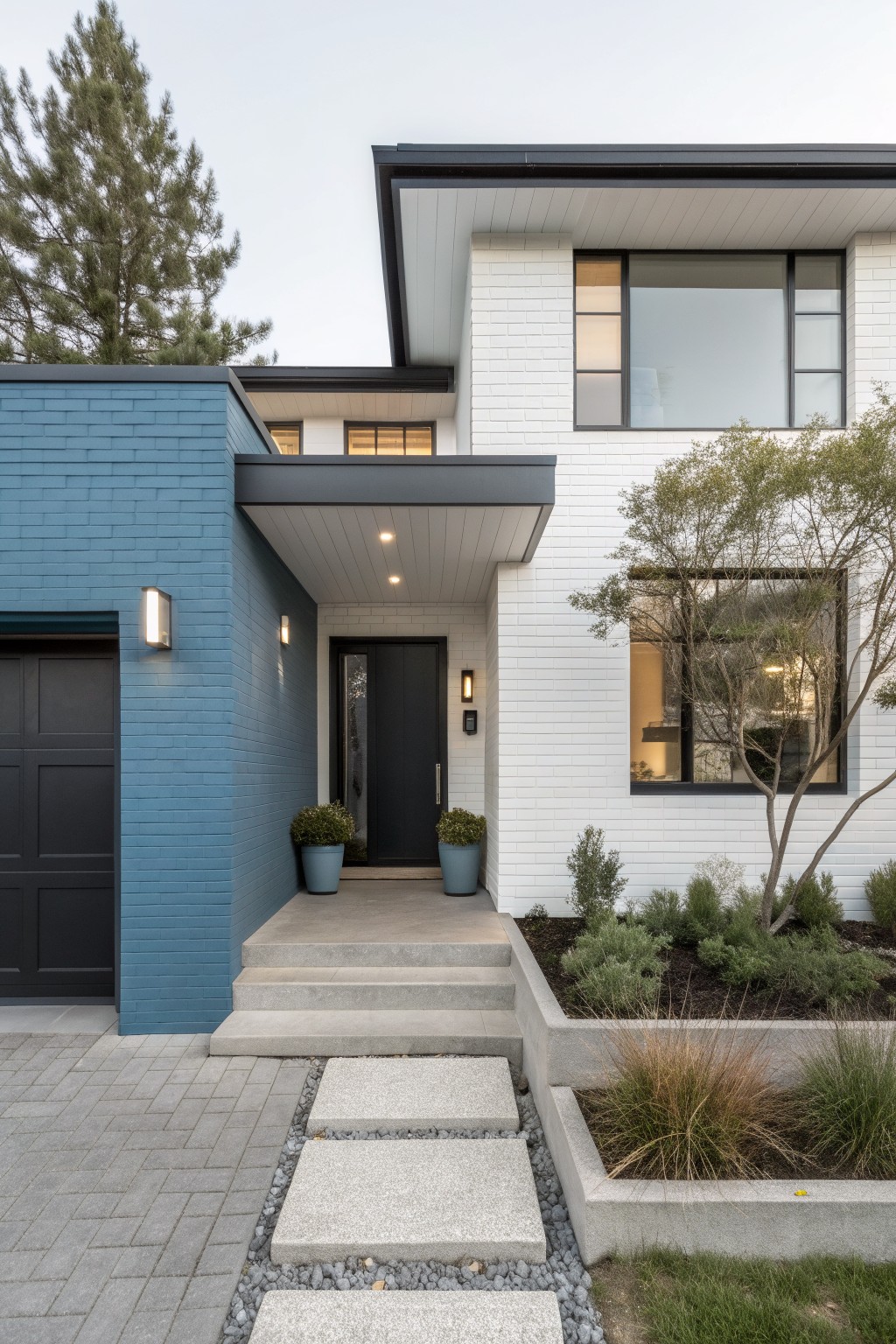 Modern house exterior with blue painted brick on the left garage section, white painted brick on the main facade, black door and window frames, concrete steps to the entry, and low landscaping with grasses and shrubs along the front.