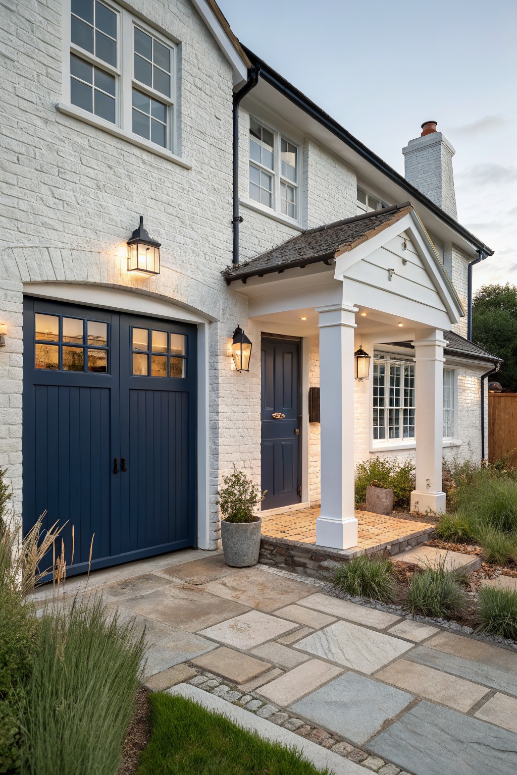 White painted brick house exterior featuring navy blue double garage doors and front door under a covered porch with columns, flanked by lanterns and a stone pathway with grasses.