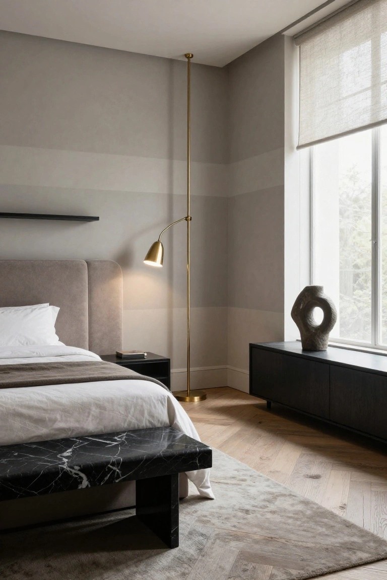 Modern bedroom featuring a bed with white linens, tall gold floor lamp, black marble bench, black sideboard with large abstract terracotta vase next to a window with sheer shades, neutral striped walls, and herringbone wood floors.