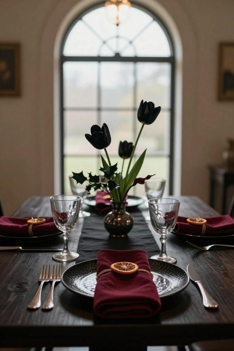 Close-up view of a dark wooden dining table set for dinner with black plates, forks and knives, red napkins tied with dried orange slices, etched wine glasses, and a centerpiece vase holding black tulips and holly branches, next to a large arched window.