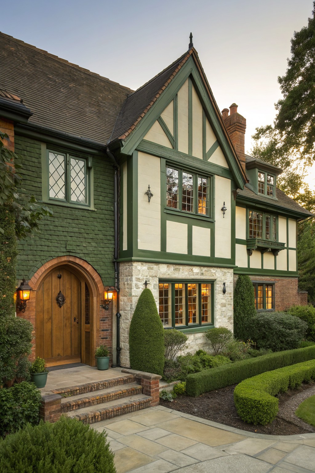 Tudor-style house exterior with dark green painted brick walls, white stucco panels, dark timber framing, arched wooden front door flanked by lanterns, stone accents, and manicured shrubs along the brick steps and pathway.