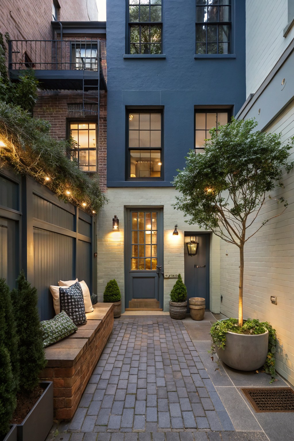 Narrow urban courtyard between brick buildings with dark navy painted walls on one side, cream brick wall with dark door on the other, central brick path, wooden bench with cushions and pillows, potted plants and topiaries, small tree in concrete pot, string lights along fence, and lanterns by the door.