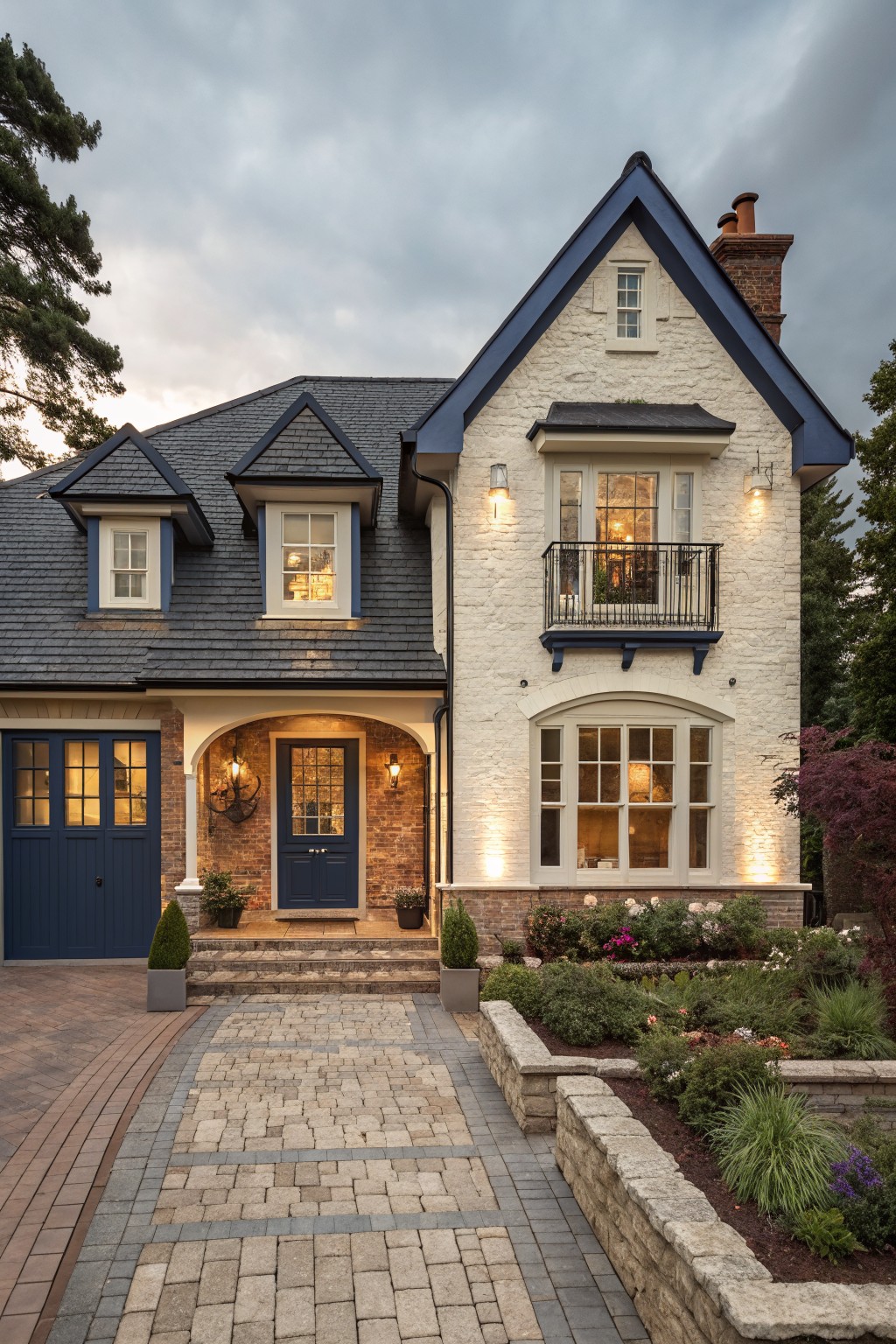 Two-story house exterior featuring white brick walls, navy blue trim on windows doors and roof edges, dark slate roof, attached garage on the left with navy doors, arched front entry, brick pavers driveway, and landscaped flower beds with plants.