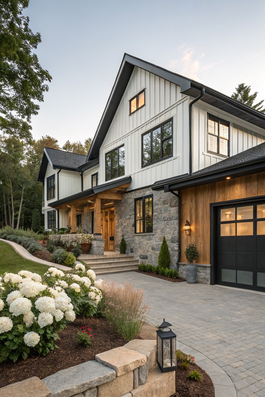 A two-story house exterior with white board-and-batten siding on the main facade, dark wood cladding on the attached garage, black roof and trim, stone entry porch, paver driveway, and landscaped front yard with white hydrangeas and low plantings.