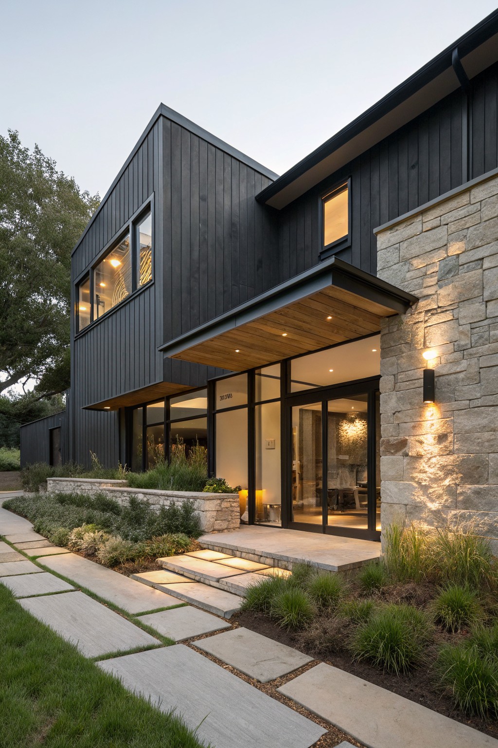 Modern house exterior with black vertical wood siding on upper walls, light stacked stone veneer on base and entry side, cantilevered wood canopy over glass pivot door, concrete slab steps and path, and low native plantings beside a lawn.