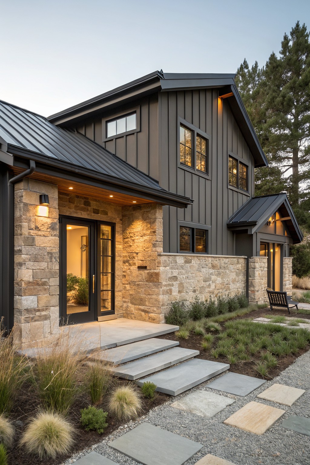 Modern farmhouse exterior with dark board-and-batten siding, stone veneer base and entry surround, metal roof, double glass doors, concrete steps, gravel path, ornamental grasses, and pine trees nearby.