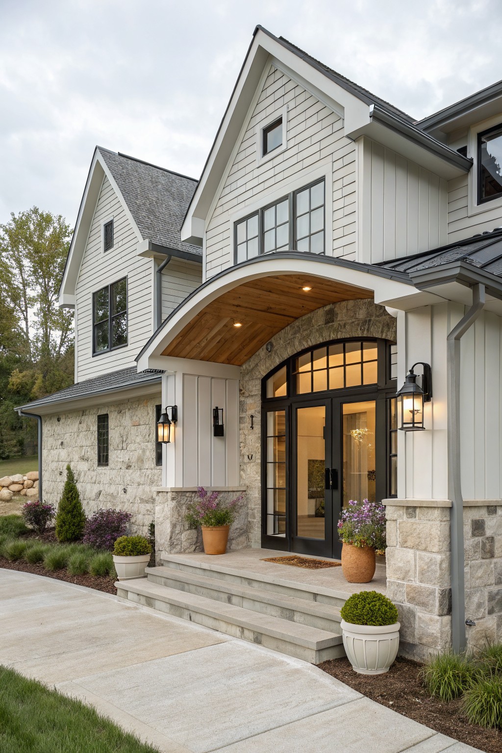 Front exterior of a two-story modern farmhouse with white shiplap siding, gray standing-seam roof, arched wooden portico over black double doors with sidelights, stone veneer on base and pillars, wall lanterns, potted plants, concrete steps, and walkway bordered by landscaping.