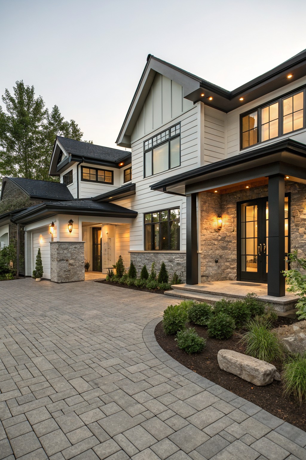Front view of a modern farmhouse house with white shiplap siding, black-framed windows and double doors, stone veneer pillars and accents at the covered entry, attached garage, paver driveway, and evergreen shrubs.