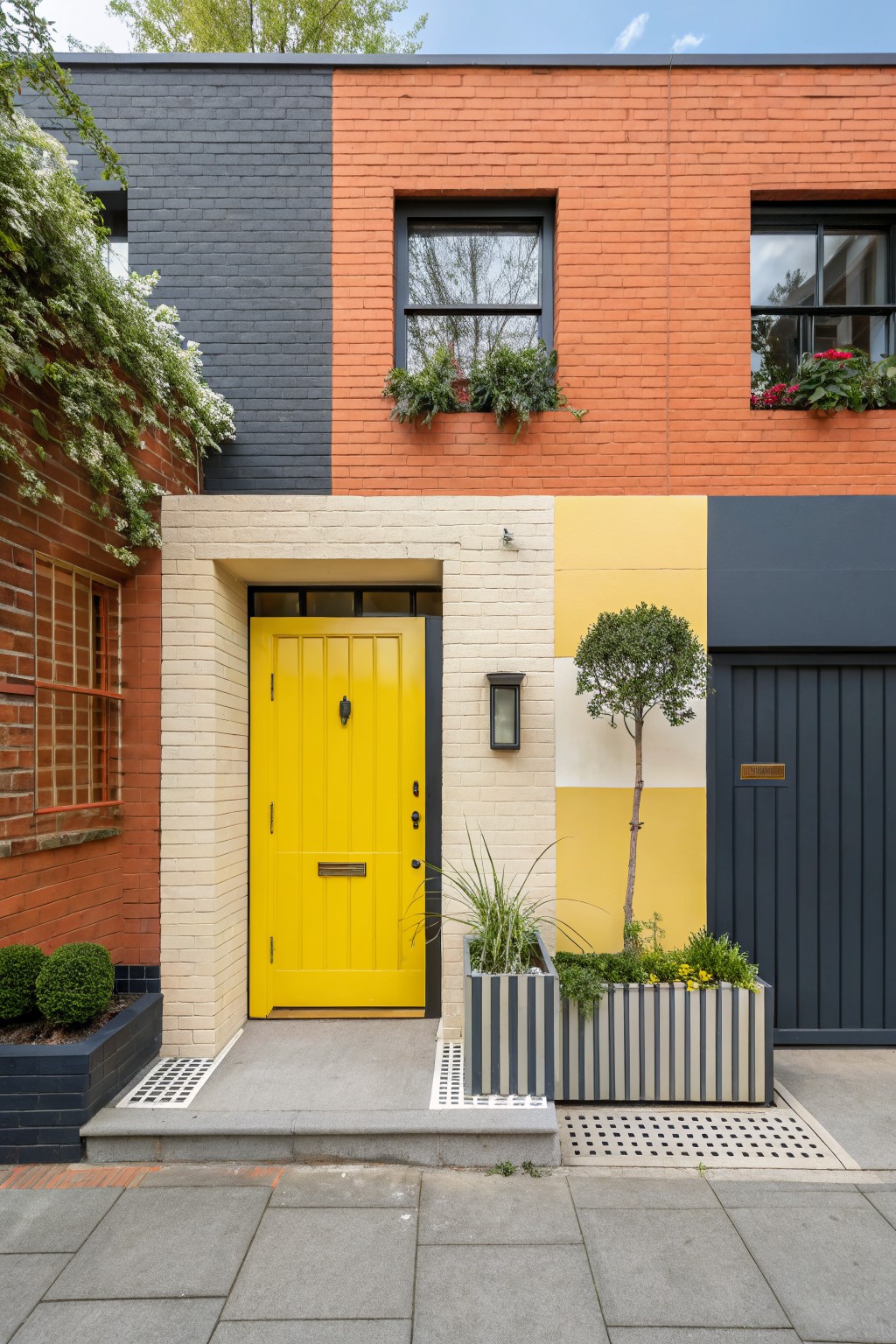 Terraced house exterior with brick painted in color blocks of dark gray, orange-red, cream, yellow, and dark gray-blue, a yellow front door, potted plants, a small tree in a yellow-painted section, and a gray garage door.