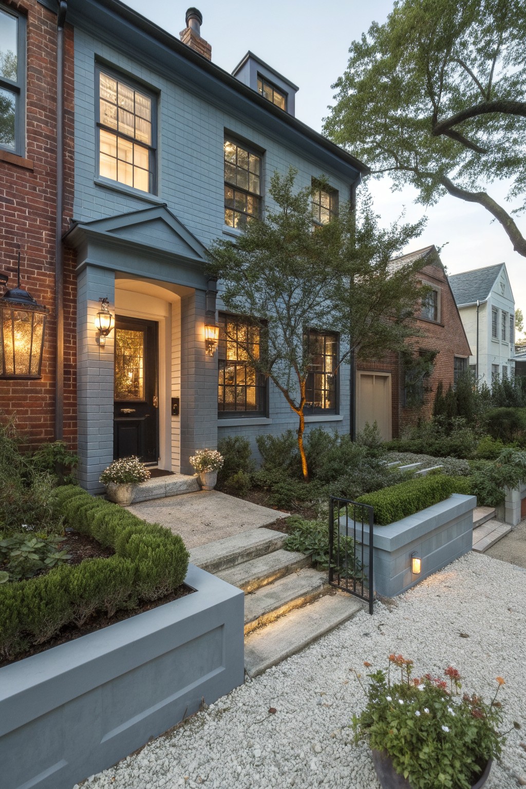Front view of a two-story house with left side in red brick and main facade painted soft blue gray, black front door under a small porch with lanterns, boxwood hedges in raised beds, gravel path, and adjacent houses visible.