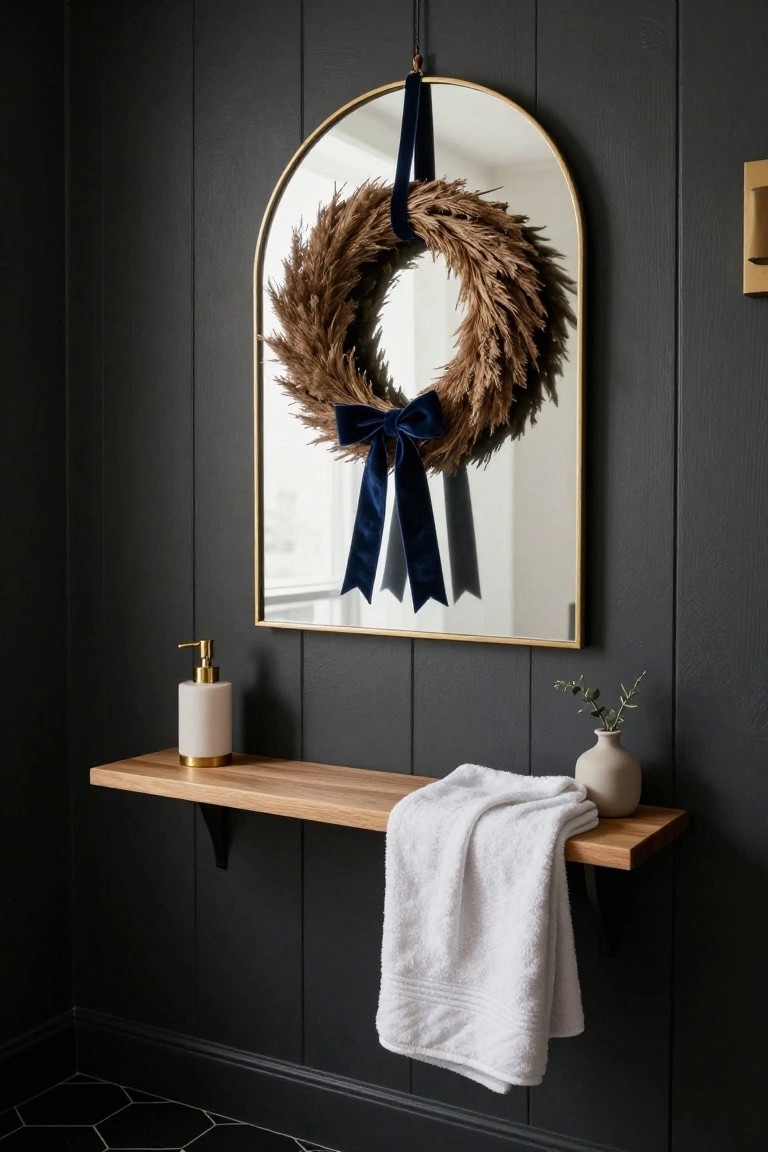 Dark shiplap wall in bathroom corner with gold arched mirror displaying wheat wreath tied with blue velvet ribbon, wooden floating shelf holding white folded towel, white ceramic vase with eucalyptus, gold soap dispenser, and black hexagonal floor tiles.