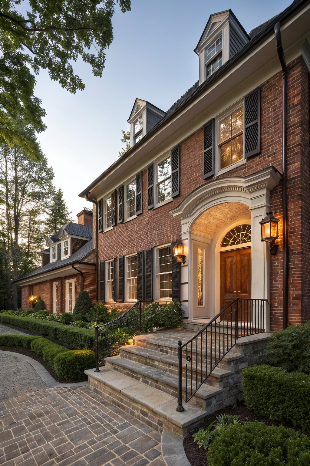 Two-story red brick house exterior with black shutters on multi-pane windows, white arched entry with wooden double doors flanked by sidelights, black lanterns, stone steps with black metal railing, boxwoods, and trees in evening light.