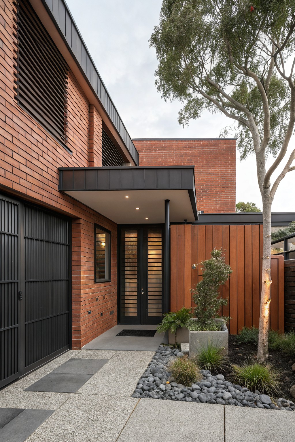 Red brick house exterior featuring black metal garage door, slatted black screens on windows and double entry doors, black metal canopy over the entrance, timber side cladding, and small landscaped entry area with plants and pebbles.
