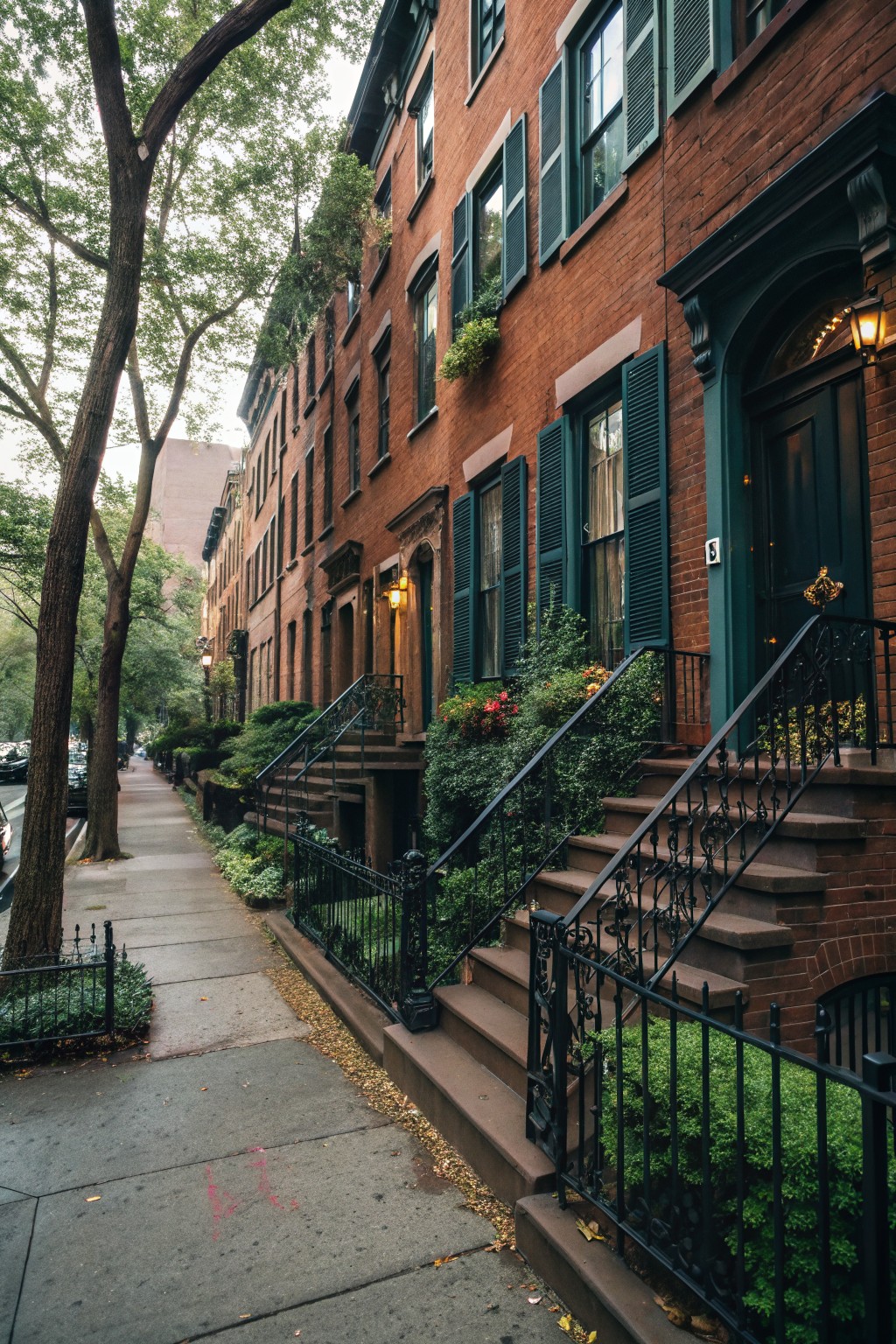 Row of four-story red brick townhouses with dark teal shutters, front steps with wrought iron railings, potted plants, and trees along a city sidewalk.