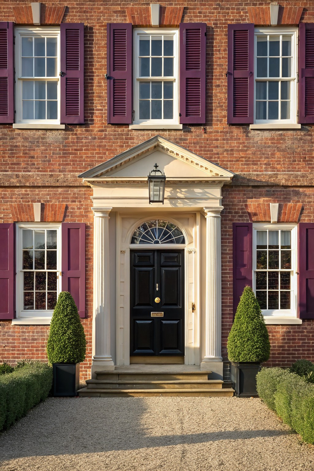 Red brick house facade with purple shutters on white-framed multi-pane windows, a white pedimented entry portico with columns framing a black front door and brass knocker, flanked by potted boxwood topiaries, gravel path, and low boxwood hedges.