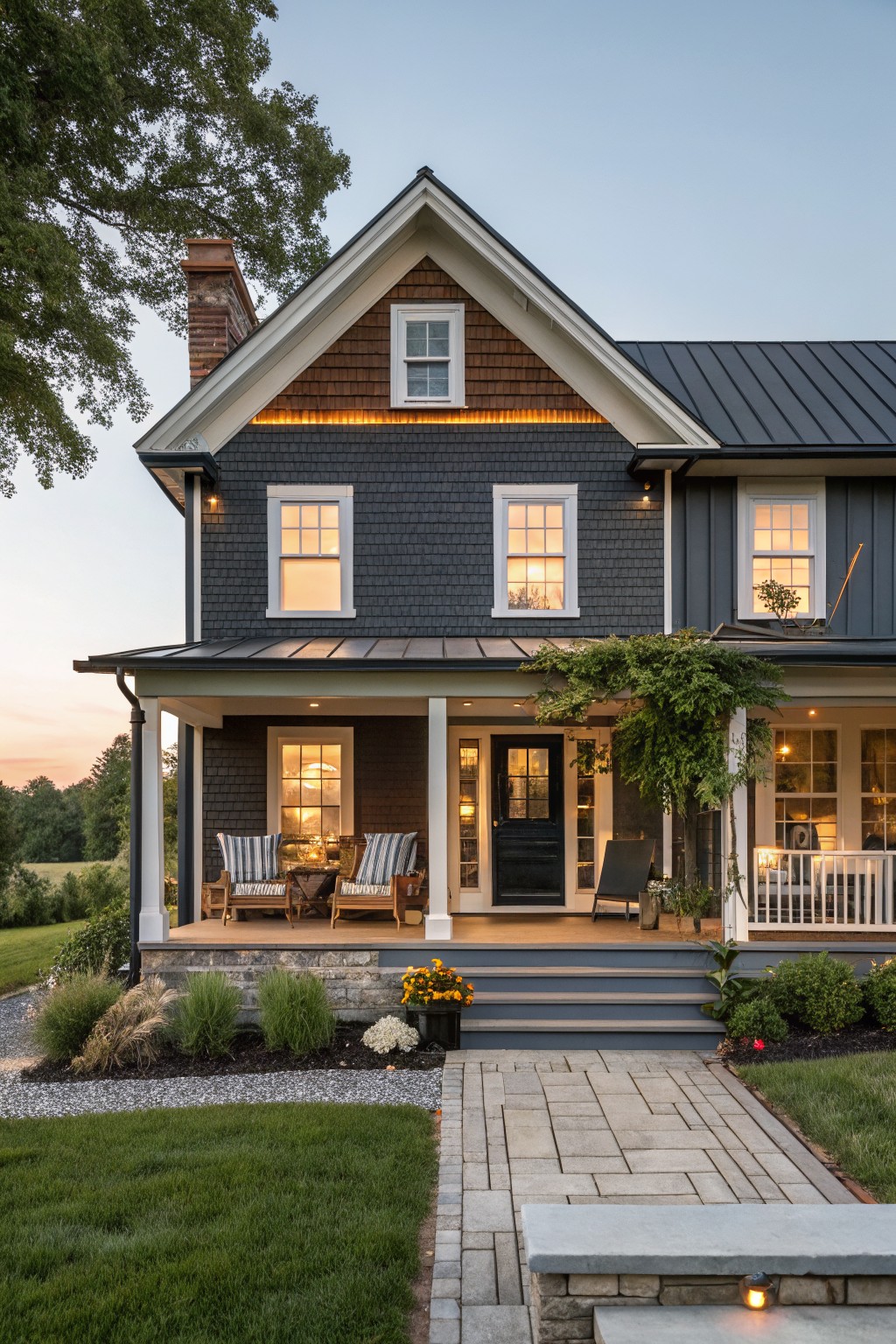 Two-story house with dark gray shingle and clapboard siding, black metal roof, brick chimney, covered porch with furniture, stone steps, and front yard landscaping at dusk.