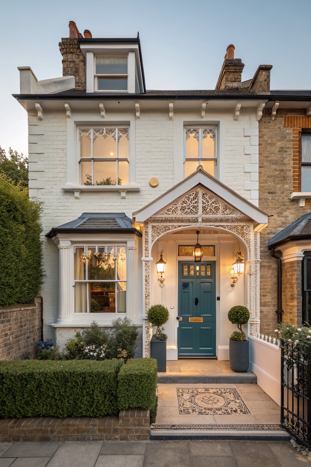 White painted brick two-story terraced house exterior with ornate porch canopy, dark green paneled front door, lanterns, topiary plants in pots, and low boxwood hedge along the front path at dusk.