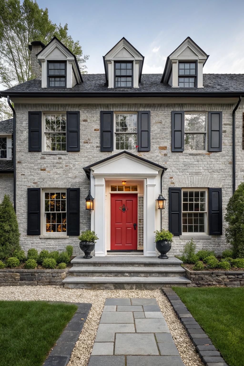 Front exterior of a two-story light gray painted brick house with black shutters, white trim around windows and dormers, red front door flanked by lanterns and large black planters, gray stone steps, bluestone walkway edged in gravel and grass.