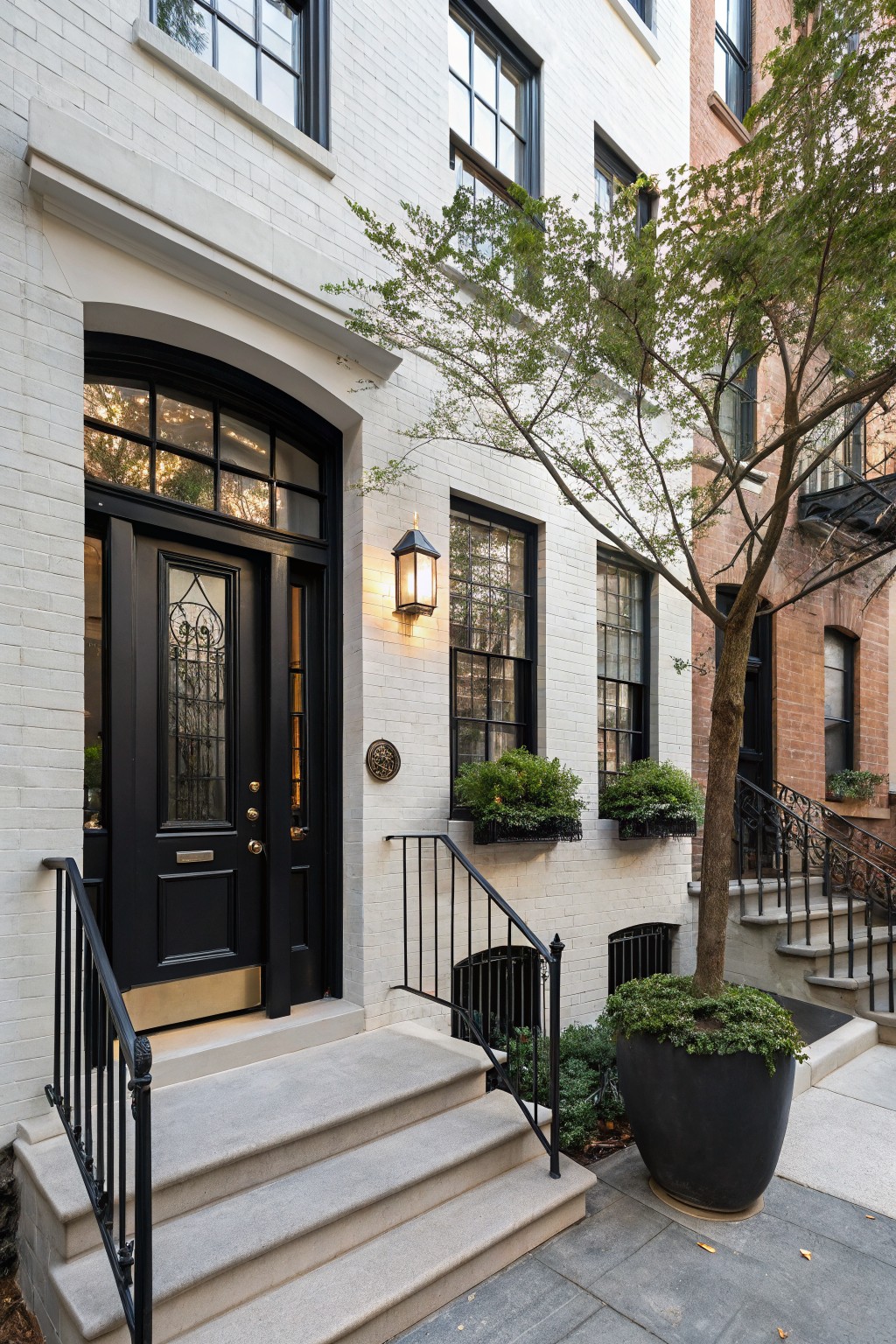 White painted brick townhouse facade with black-framed windows, arched black front door, stone steps with black railings, wall-mounted lantern, and greenery in planters beside a tree on a city sidewalk.