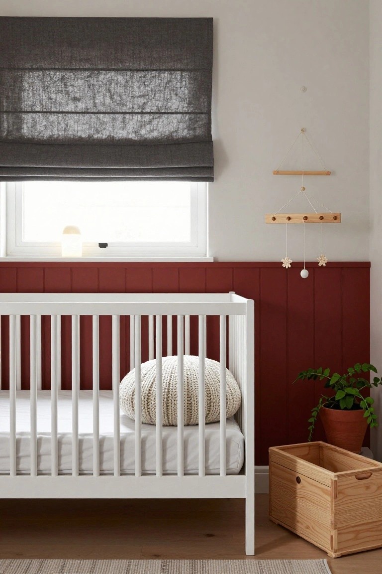 White crib in a nursery with cherry red vertical paneled lower walls, gray fabric blinds on the window, wooden mobile overhead, knit pillow on the bed, potted plant, and wooden storage box nearby.