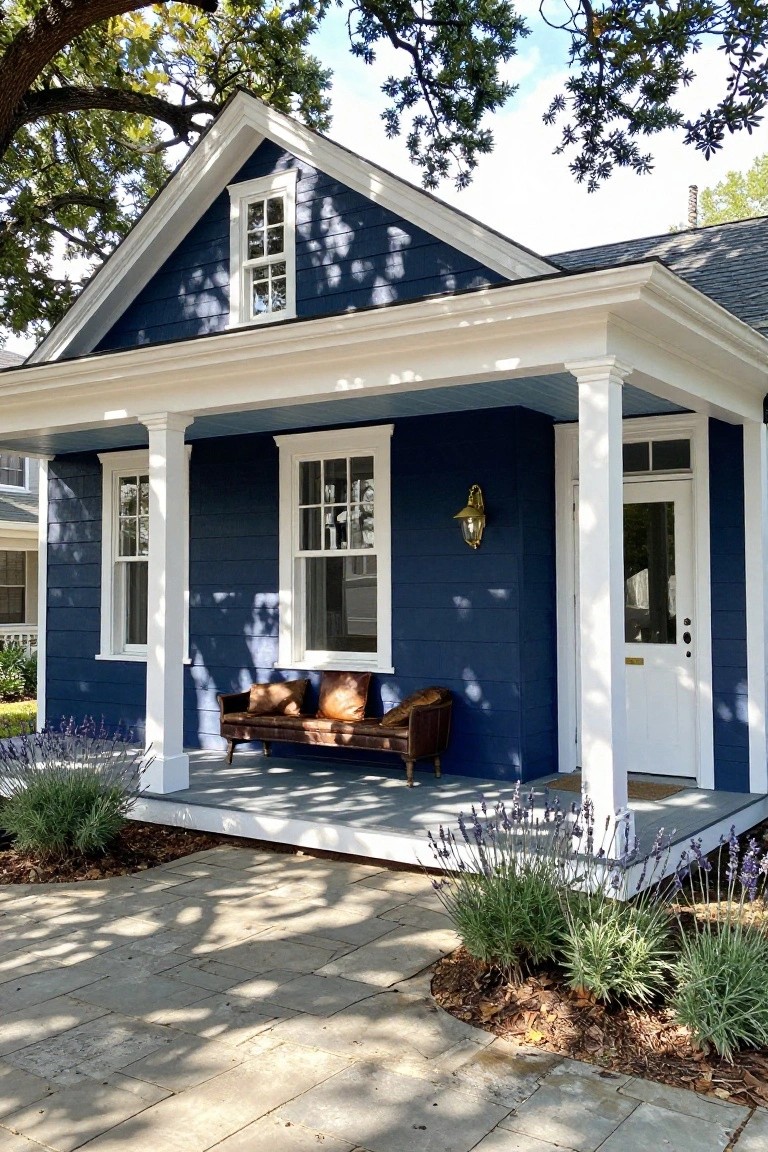 Navy blue colonial house with white trim, gabled roof, front porch supported by columns, leather bench swing, gold wall lantern, white door, and lavender plants along a stone pathway.