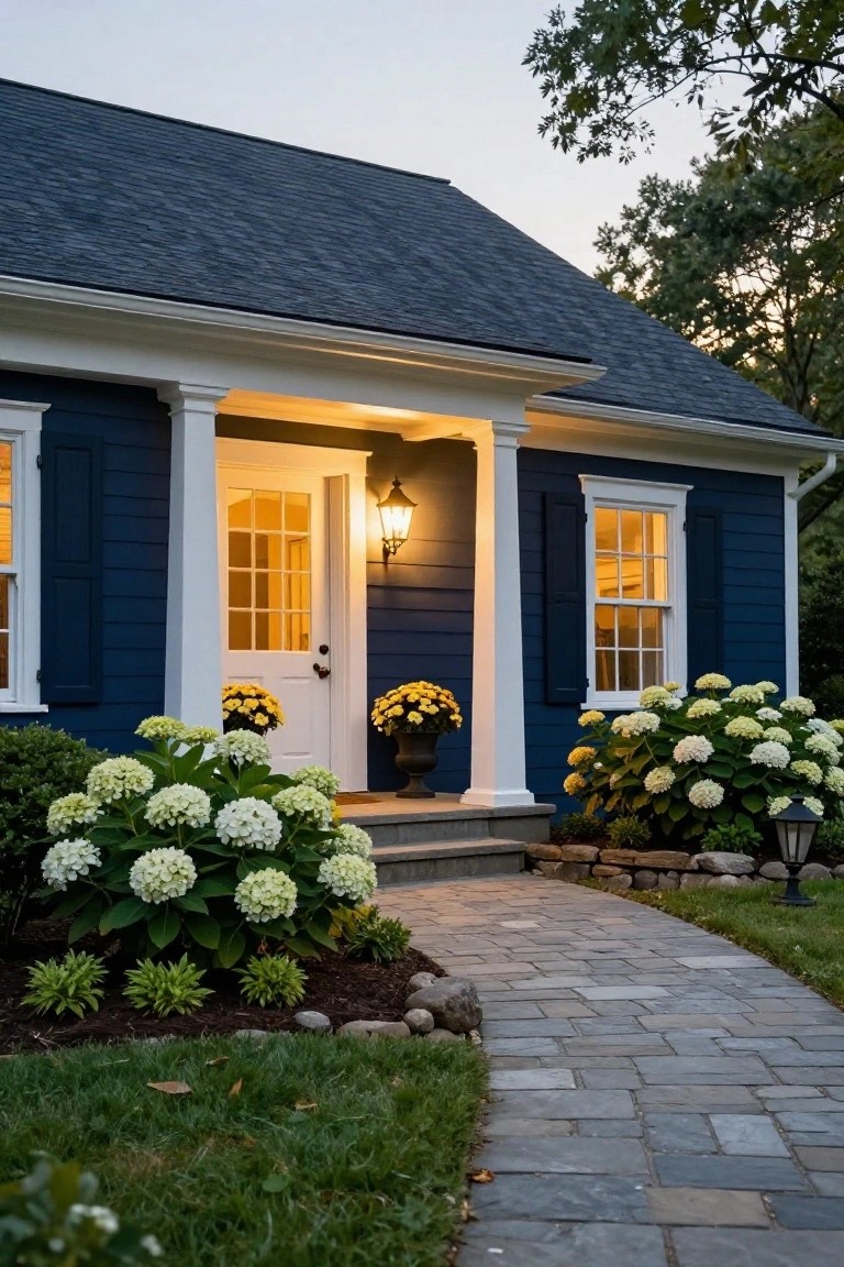 Front exterior of a navy blue clapboard colonial house with white trim, columns, open porch door, lanterns, hydrangeas, and stone pathway at dusk.