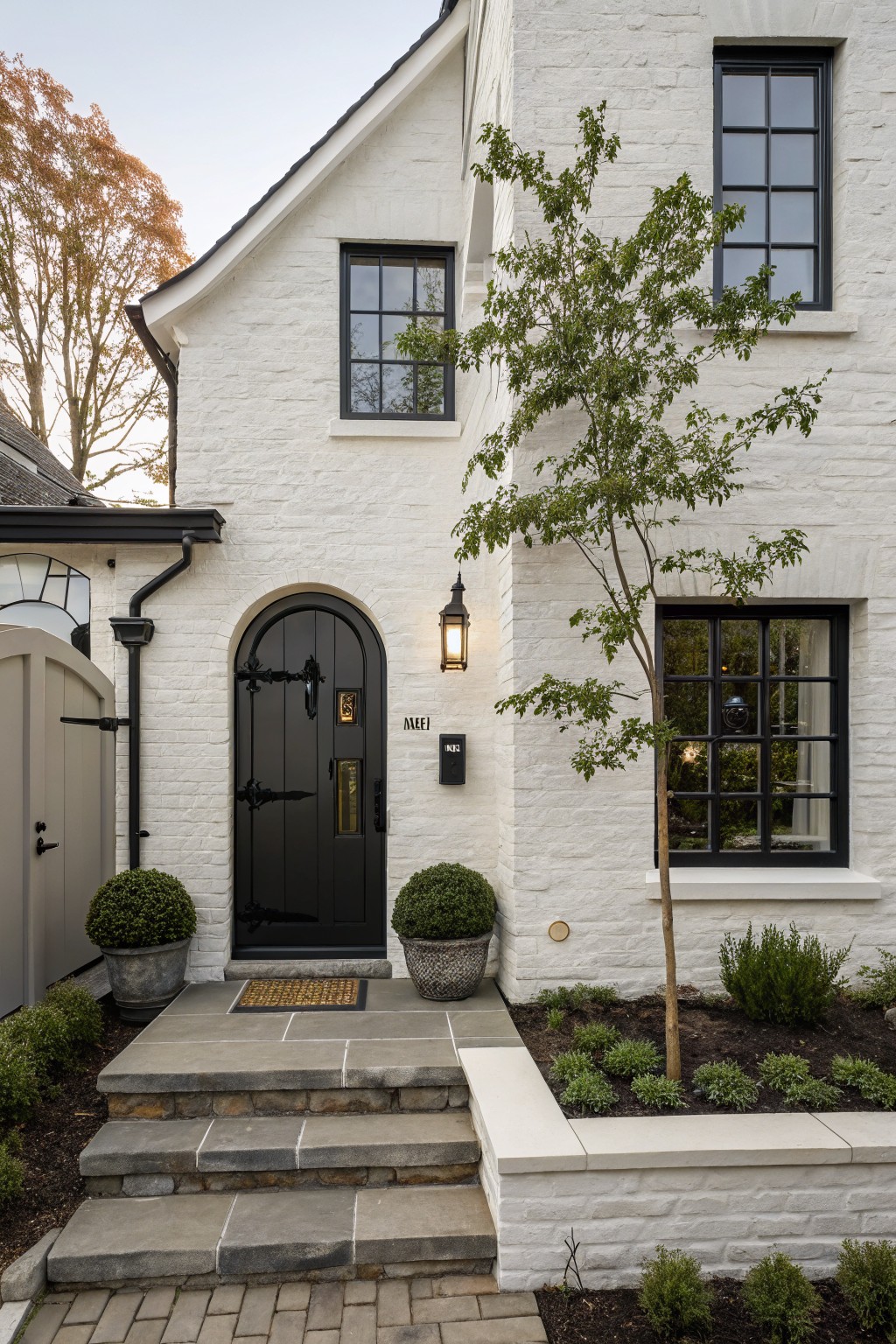 White painted brick house exterior with black arched front door, black-framed windows, stone entry steps, potted topiaries, wall lantern lights, mailbox, and small tree in planting bed beside the door.